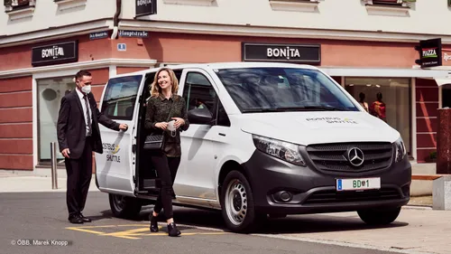 Eine Frau lächelt, während sie aus einem weißen Mercedes-Van mit der Aufschrift Postbus Shuttle aussteigt. Ein Mann hält die offene Tür hinter ihr.