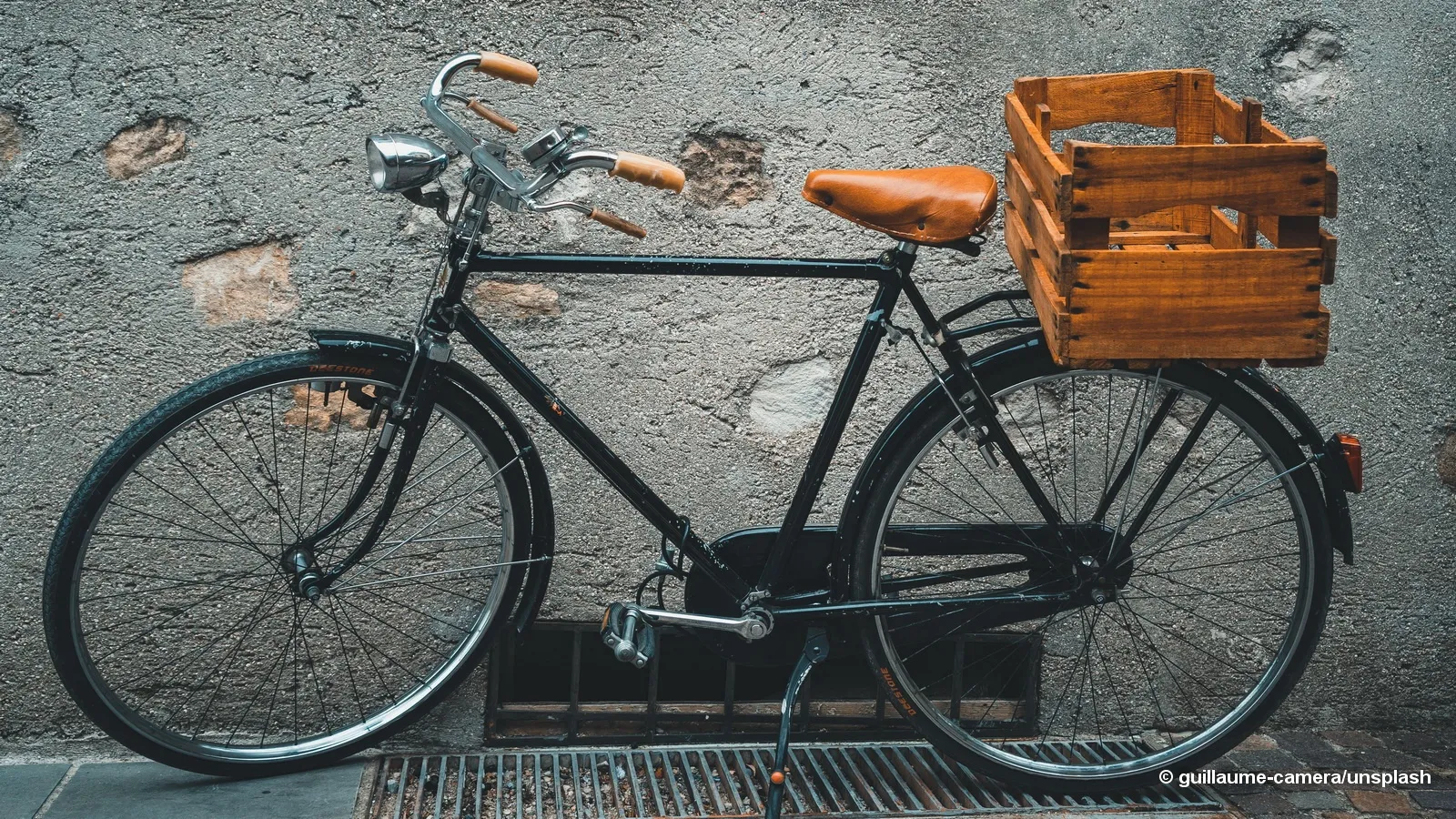 Ein schwarzes Fahrrad mit braunen Lenkern und einem braunen Ledersitz steht auf einer Betonfläche mit einem hölzernen Kasten hinten.