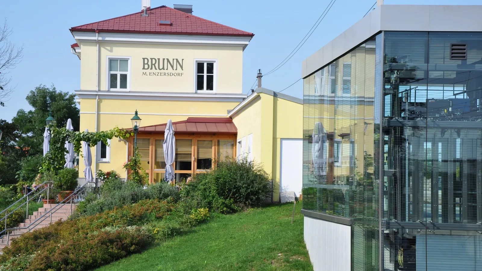 Das Äußere von Brunn M. Enzersdorf zeigt ein gelbes Gebäude mit rotem Dach, Glasfenstern und einem Garten davor. Vor dem Eingang steht ein geschlossener Regenschirm.