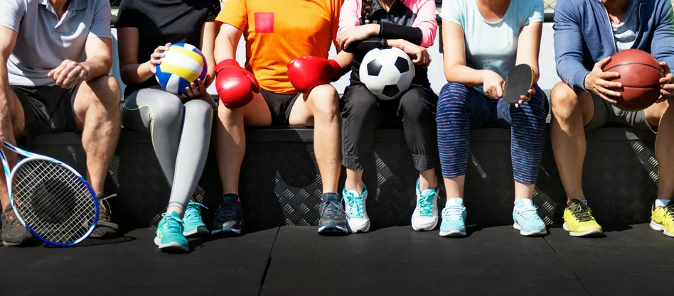 Vier Personen sitzen auf einer Bank, tragen Sportkleidung und halten Sportgeräte. Einer hat einen Fußball, ein anderer einen Volleyball und zwei haben Boxhandschuhe.
