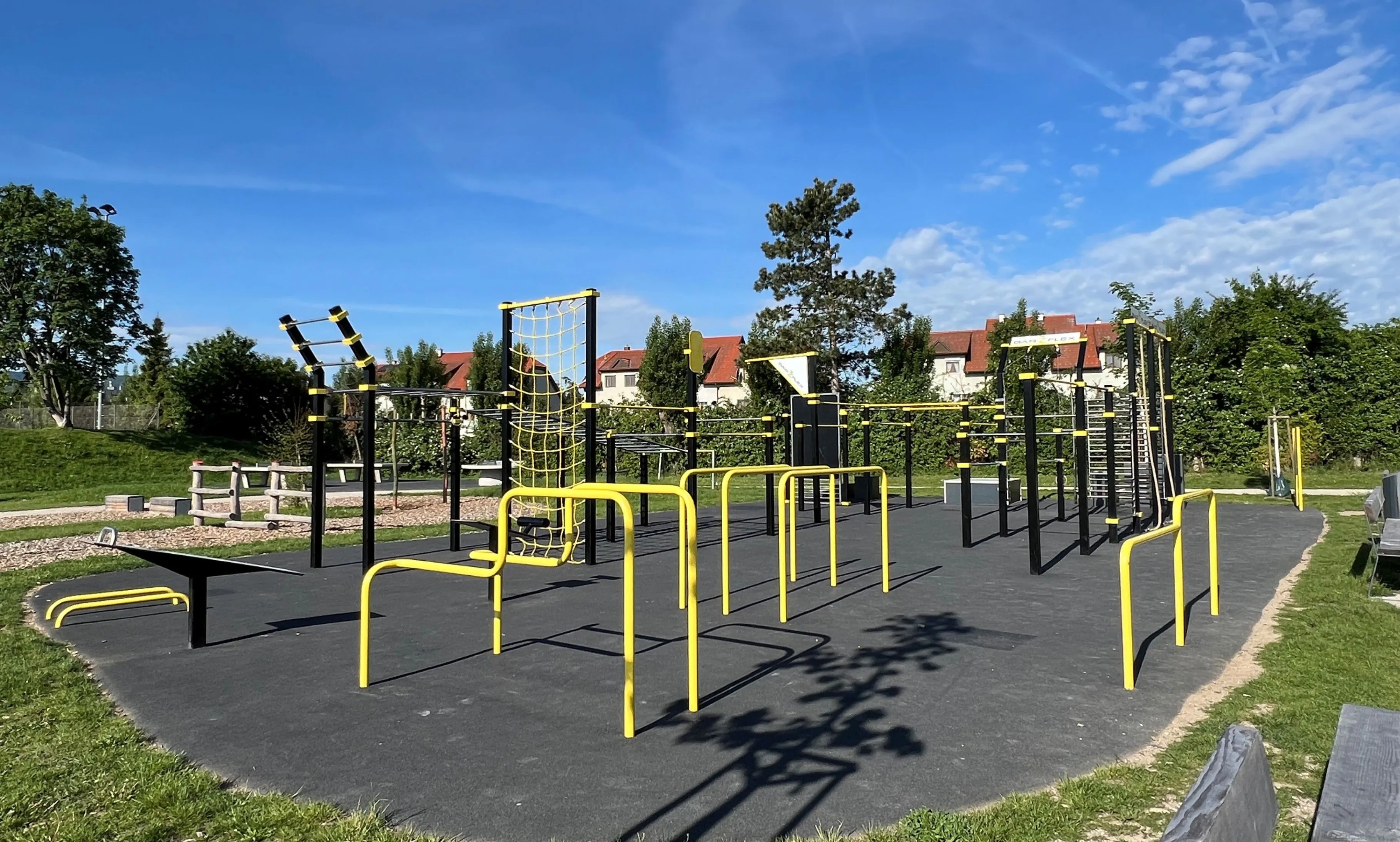 Ein Outdoor-Fitnesspark mit gelben und schwarzen Geräten, umgeben von Bäumen und Häusern. Der Park ist vom blauen Himmel gut beleuchtet.