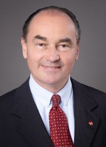 Ein lächelnder Mann in Anzug und roter Krawatte, mit einem kleinen Logo auf seiner Jacke, steht vor einem grauen Hintergrund.