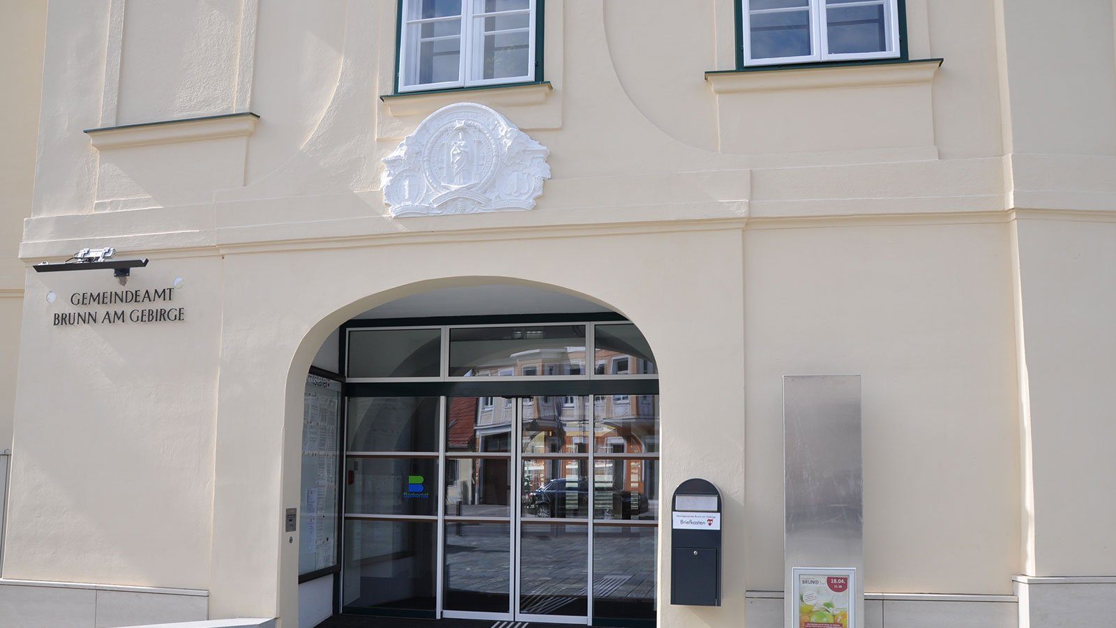 Ein beigefarbenes Gebäude verfügt über einen weißen, gewölbten Eingang mit Glastüren und Fenstern sowie einem dekorativen Emblem über dem Eingang. Ein schwarzer Briefkasten ist an der Wand montiert.
