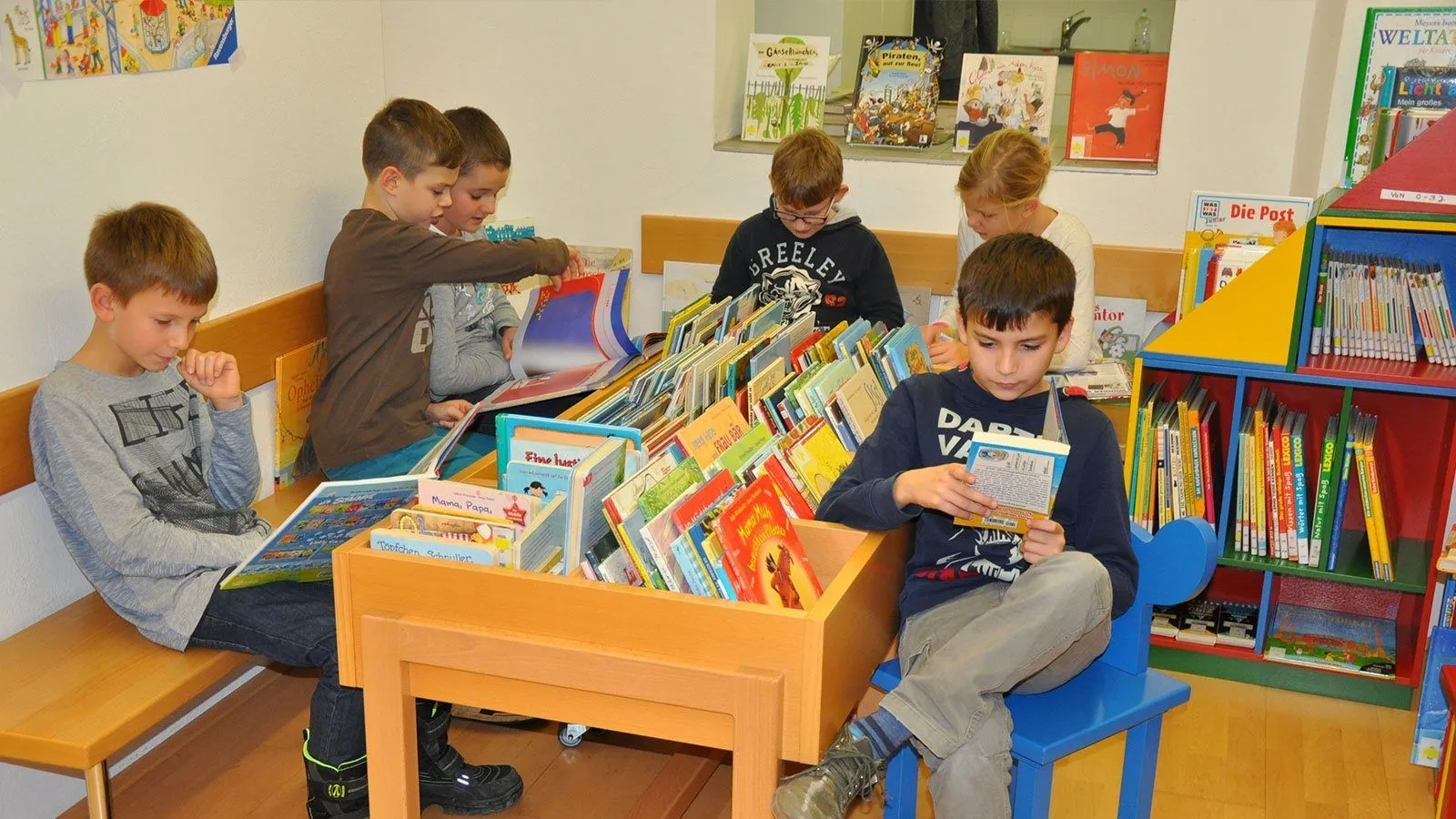 Kinder sitzen in einer Bibliothek und stöbern in Büchern. Ein Junge liest, während ein anderer auf ein Buch zeigt. Ein Mädchen sitzt neben einem Jungen und schaut Bücher an.