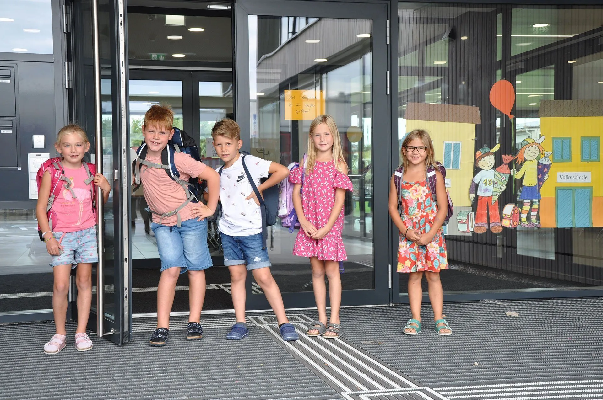 Vier Kinder stehen lächelnd vor einem Gebäudeeingang mit Rucksäcken. Ein gelbes Schild ist im Hintergrund sichtbar.