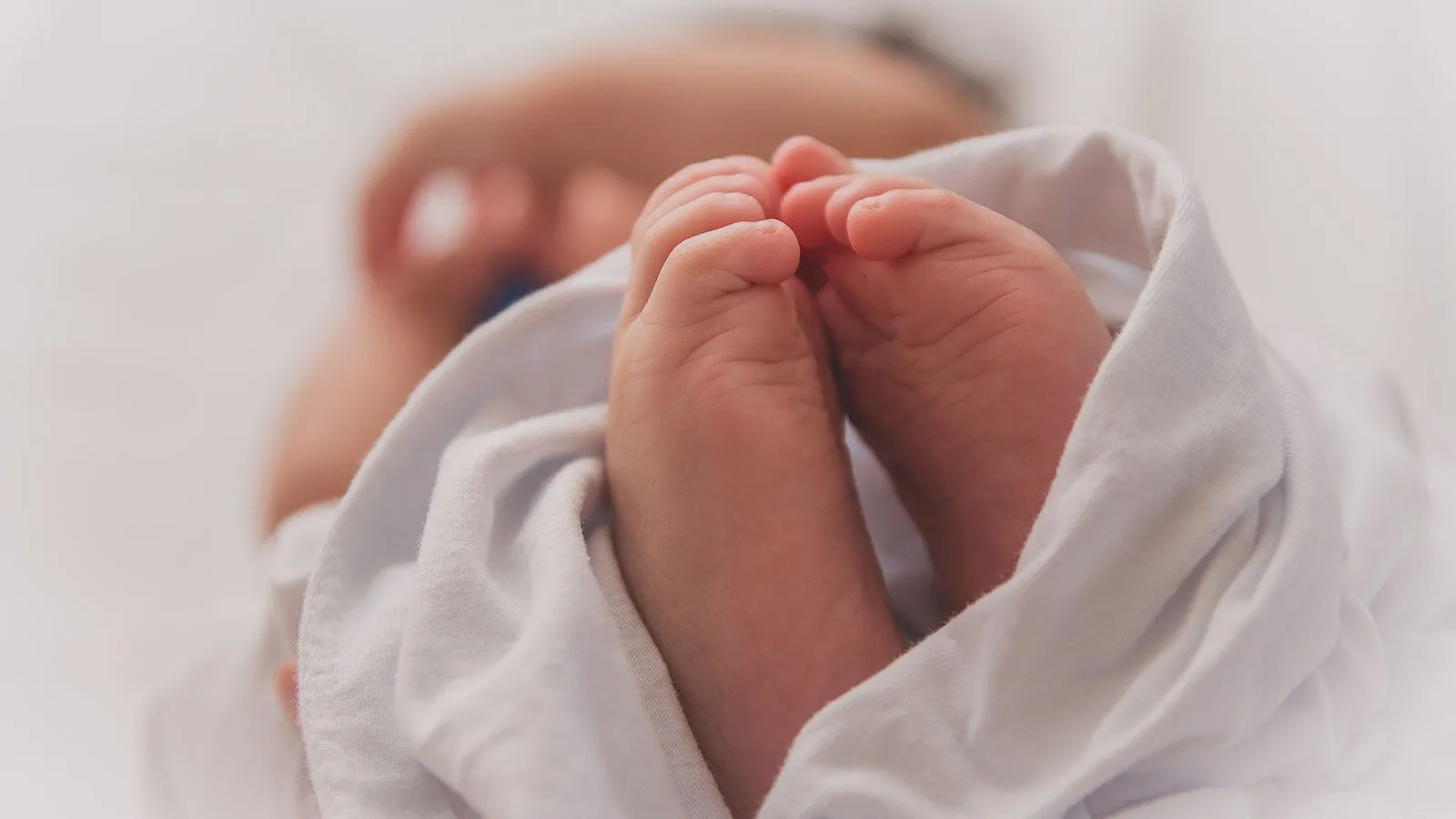 Nahaufnahme der Füße eines Neugeborenen, das in eine weiße Decke gewickelt ist, wobei der Rest des Babys im Hintergrund unscharf ist.