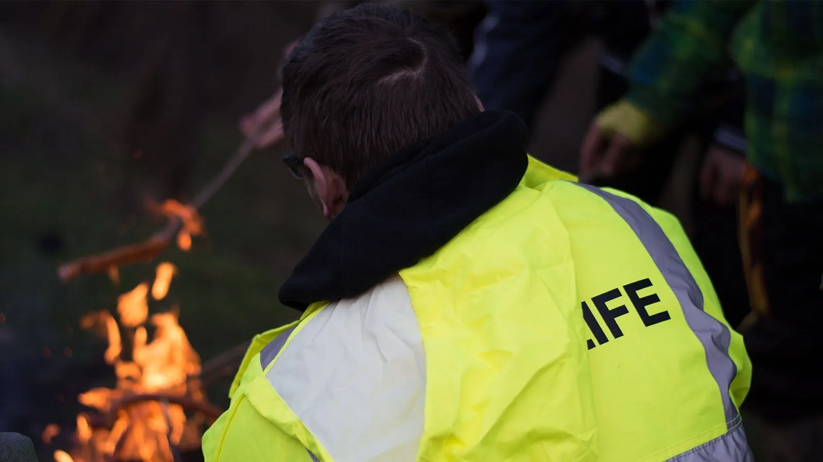 Ein Mann in einer neongelben Sicherheitsweste mit dem Wort 'IFF' darauf blickt in einem dunklen Bereich nach unten. Es gibt eine verschwommene Person und ein Feuer im Hintergrund.