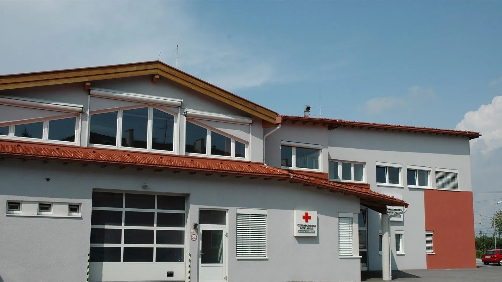 Ein weißes Gebäude mit rotem Dach hat mehrere Glasfenster und ein rotes Kreuz-Schild an der Wand. Das Schild ist mit "Rettungswache" und "Notruf" beschriftet. Das Gebäude hat eine weiße Tür mit Glasfenster und eine Garagentür.