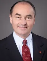 Ein Mann mit grauem Haar trägt einen dunklen Anzug, ein weißes Hemd und eine rote Krawatte mit kleinen Punkten und einem kleinen Logo an der Revers.