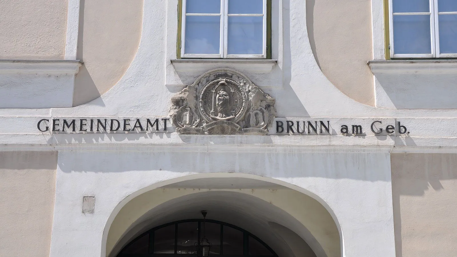 Ein weißes Gebäude zeigt ein Fenster und ein Schild über einem gewölbten Eingang. Das Schild steht 'NDEAMT BRUNN a' mit einem Wappen darüber.