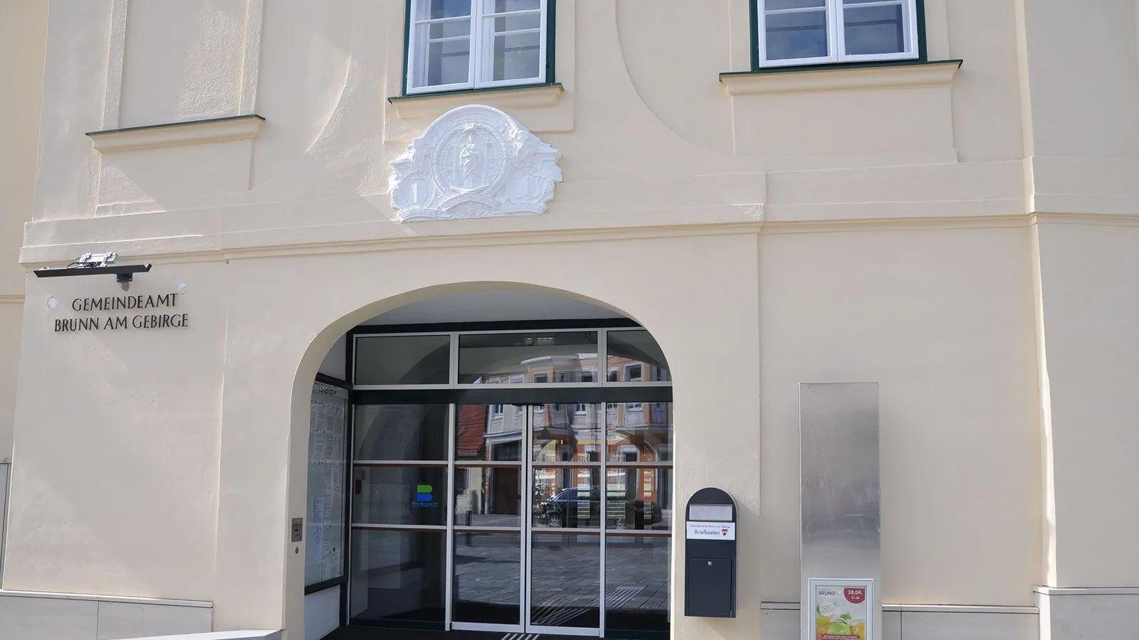Das Äußere eines cremefarbenen Gebäudes mit einem gläsernen Bogeneingang, einem Schild und einem gewölbten Wandteil über dem Eingang.