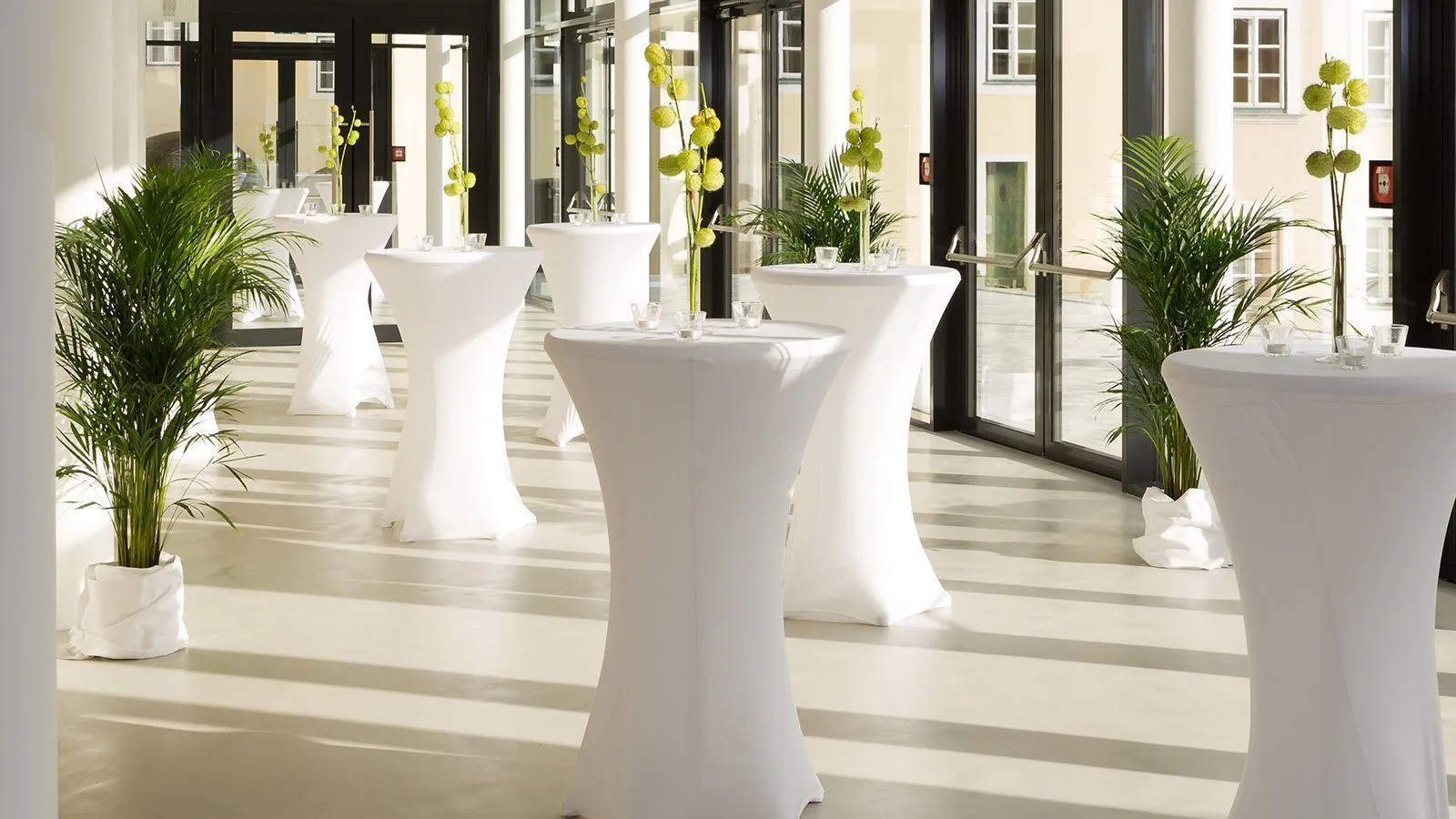 Ein moderner Raum mit weißen Tischen und Stühlen, ausgestattet mit gelben Blumenarrangements in Glasvasen. Der Raum hat große Glastüren und einen glänzenden Boden.