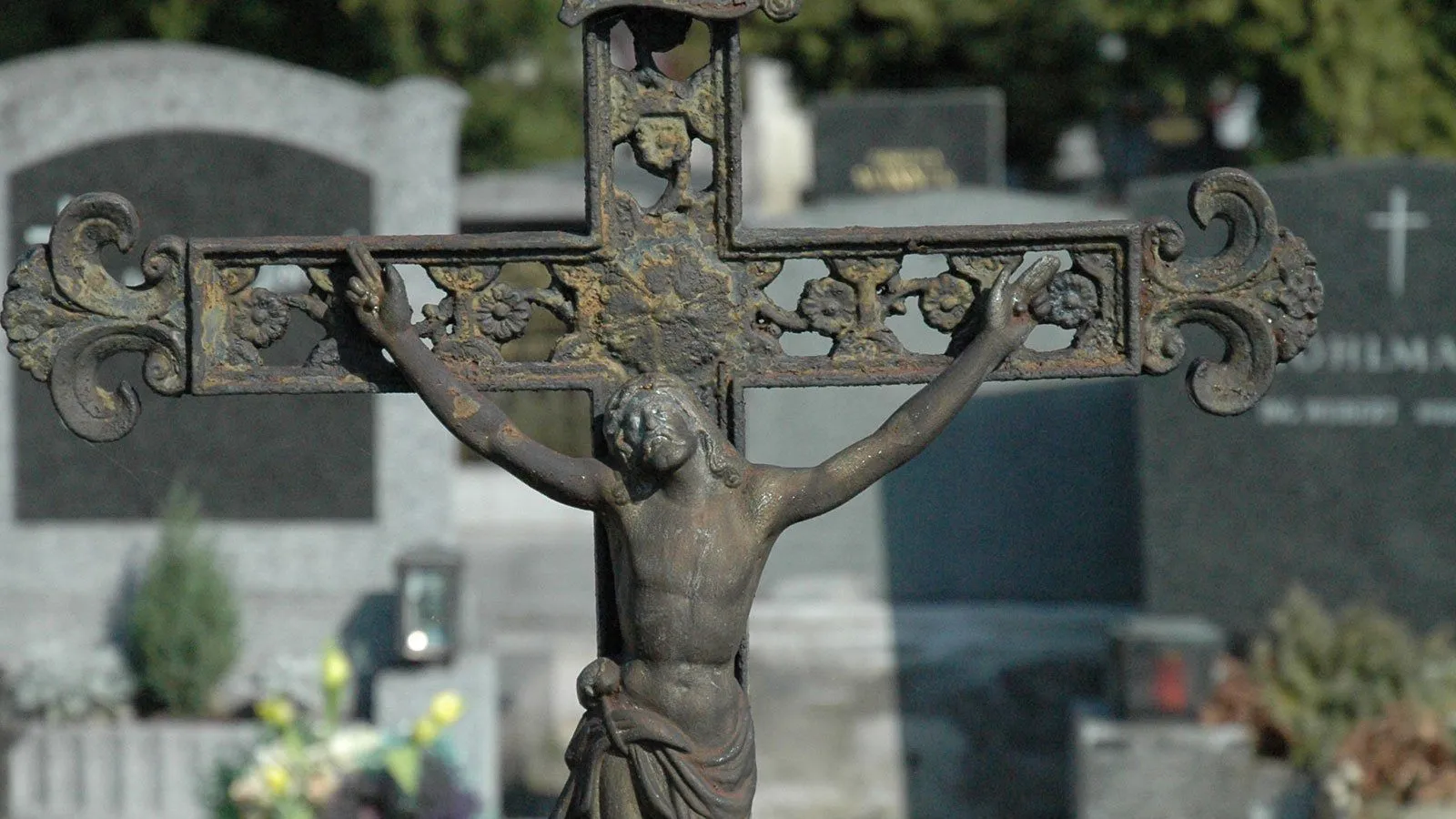 Ein verwittertes Bronzekreuz steht auf einem Friedhof, verziert mit Blumenmustern und Rost. Die Figur Christi ist an einem vertikalen Kreuz mit ausgestreckten Armen befestigt.