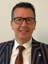 Ein Mann mit Brille, Anzug und Krawatte lächelt in die Kamera. Er hat kurze Haare und trägt eine Anstecknadel auf der Brust.