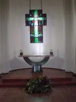 Ein modernes, farbiges Kreuz hängt über einem Altar in einer ruhigen Kirche. Der Altar ist schlicht mit zwei Kerzen auf beiden Seiten. Eine Pflanze steht vor dem Altar auf dem Boden.