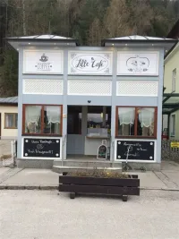 Ein kleines Café mit weißer Außenfassade und grünem Dach. Es hat ein Schild mit der Aufschrift 'COFFEE' und 'Lille Cafe'. Es gibt Kreidetafeln mit Werbung für frisches Essen und hausgemachte Desserts.