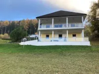 Ein zweistöckiges Haus mit schwarzem Dach steht auf einem Hügel, umgeben von grünem Gras und Bäumen. Ein weißes Auto ist in der Nähe geparkt.