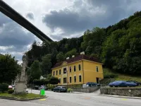 Ein gelbes Haus mit einem braunen Dach und mehreren Fenstern steht nahe einer Straße. Eine Brücke spannt sich darüber, und Autos sind davor geparkt.