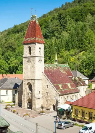 Eine mittelalterliche Kirche mit einem roten Dach steht in einer kleinen Stadt. Sie hat einen Turm mit einer Uhr, umgeben von einem üppigen, grünen Hang.