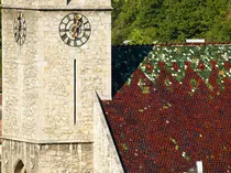 Eine Uhr an einem Steinturm zeigt die Zeit an. Das Dach ist mit bunten Fliesen bedeckt. Bäume sind im Hintergrund sichtbar.