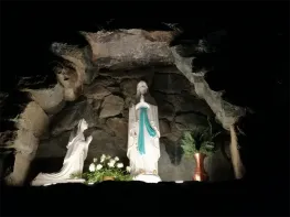 Eine weiße Statue einer Frau befindet sich in einer Höhle mit einer anderen Statue und einem Blumenvase. Die Statue hat eine blaue Schärpe und ein Kreuz am Hals.