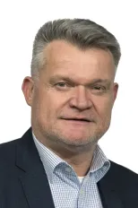 Ein Mann mit grauem Haar trägt einen schwarzen Anzug und ein blaues kariertes Hemd und lächelt in die Kamera, vor weißem Hintergrund isoliert.