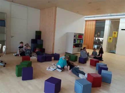 Eine Gruppe von Kindern und Erwachsenen sitzt auf mehrfarbigen Kissen in einem Raum, liest Bücher und interagiert miteinander. Der Raum hat Holzböden, weiße Wände und einen Holzschrank mit Büchern.