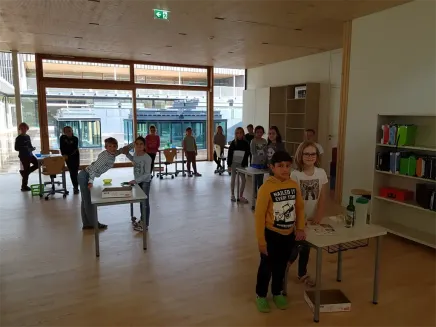 Eine Gruppe von Kindern in einem Klassenzimmer mit Tischen und Stühlen, einige führen Aktivitäten durch. Es gibt Glasfenster und Türen sowie ein Regal mit Gegenständen.