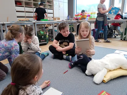 Kinder sitzen auf dem Boden in einem Klassenzimmer, einige mit Büchern, eine mit einem Tablet. Ein Mädchen hält ein Plüschtier, andere schauen zu. Ein Lehrer steht an einem Tisch im Hintergrund.