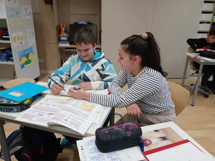 Ein Junge und ein Mädchen sitzen an einem Schreibtisch und halten jeweils einen Stift. Sie arbeiten an einem offenen Buch und Papier. Hinter ihnen sind ein Computer und ein Bücherregal zu sehen.