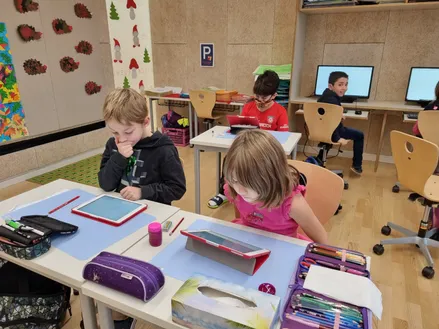 Vier Kinder sitzen in einem Klassenzimmer an Schreibtischen mit Tablets und Büchern. Ein Junge trägt eine Gesichtsmaske. Hinter ihnen benutzt ein Junge einen Computer. Verschiedene Schulsachen liegen auf den Schreibtischen.