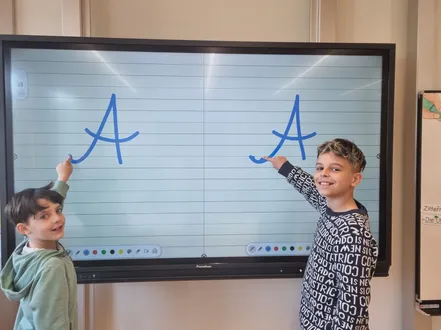 Zwei junge Jungen stehen vor einer großen Tafel. Ein Junge zeigt auf den Buchstaben A auf der linken Seite und der andere auf den Buchstaben A auf der rechten Seite. Die Tafel hat eine blaue Linie oben und unten.