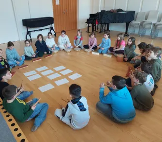 Eine Gruppe von Kindern sitzt kreisförmig auf dem Boden und spielt Trommeln und andere Musikinstrumente. Ein Klavier steht hinter ihnen.