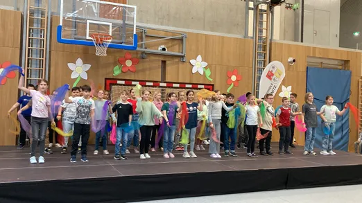 Eine Gruppe von Kindern auf der Bühne mit bunten Bändern, in einer Turnhalle mit einem Basketballkorb und Blumendekorationen an der Wand.