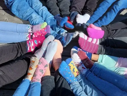 Eine Gruppe von Menschen sitzt im Kreis und trägt bunte Socken. Jede Person hat einzigartige Designs auf ihren Socken und sie sitzen auf einer Holzoberfläche.
