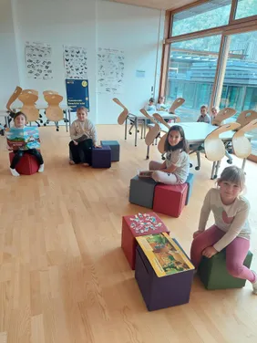 Kinder sitzen auf bunten Würfeln und lesen Bücher in einem hellen Klassenzimmer mit großen Fenstern. Ein Puzzle liegt auf einem roten Würfel vor ihnen.