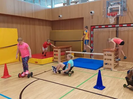 Mehrere Kinder in einer Turnhalle sind in einer körperlichen Aktivität engagiert. Ein Mädchen fährt einen Roller, eine andere sitzt auf einem Stuhl mit Rädern, während andere rennen und springen. Ausrüstung wie Matten und Kegel sind aufgestellt.