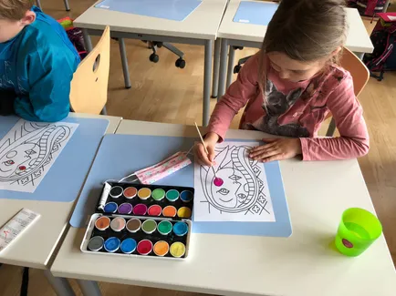 Ein junges Mädchen malt in einem Klassenzimmer ein Bild mit einem Farbpalette auf dem Tisch vor ihr. Sie trägt ein rosa Langarmshirt mit einem Känguru-Design.