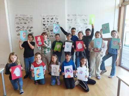Eine Gruppe von Kindern in einem Klassenzimmer posiert mit ihren Zeichnungen. Sie halten verschiedene bunte Zeichnungen hoch, die wahrscheinlich Tiere darstellen. An den Wänden im Hintergrund befinden sich pädagogische Poster.