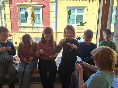 Fünf Kinder sitzen auf einer Bank und sind mit einer Strickarbeit beschäftigt. Sie sitzen vor einem Fenster mit Dekorationen. Das Gebäude hinter ihnen hat gelbe Wände und Glasfenster.