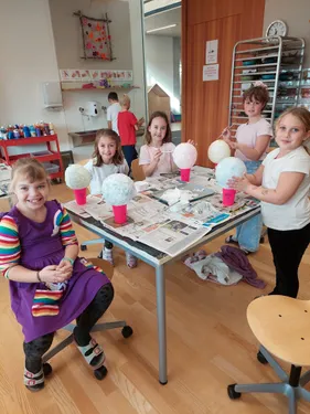 Fünf Mädchen arbeiten an Papiermaché-Projekten an einem Tisch. Eine hält ein fertiges Objekt. Andere haben halbfertige Kreationen. Sie sind in einem Raum mit einem Waschbecken, Regalen und einer Vitrine.