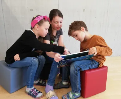Drei Kinder sitzen auf dem Boden, zwei Mädchen und ein Junge, und schauen in ein Buch. Das Mädchen links zeigt auf das Buch, während der Junge rechts liest. Sie tragen Freizeitkleidung.