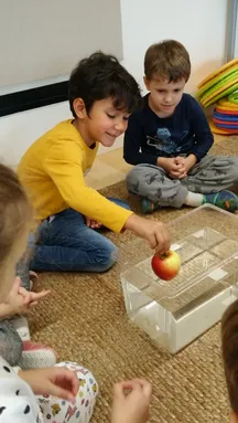 Zwei junge Jungen sitzen auf einem Teppich, einer hält einen Apfel in einem durchsichtigen Behälter. Der Junge in Blau hat ein Dinosaurier-Design auf seinem Pyjama.