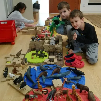 Drei Kinder spielen mit einer großen Sammlung von Tierfiguren und Bauklötzen auf einem Holzboden. Ein Mädchen sitzt links, während zwei Jungen rechts sitzen, einer von ihnen hält ein grünes Spielzeug.