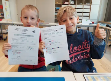Zwei Jungen halten Mathearbeitsblätter in einem Klassenzimmer hoch. Einer trägt ein rotes Shirt und der andere eine blaue Hoodie mit 'Velocity Industries' darauf. Sie lächeln beide.
