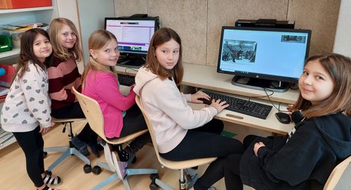 Zwei Mädchen sitzen in einem Klassenzimmer an Computern, eine benutzt die Tastatur, während die andere in die Kamera lächelt. Dahinter zeigt ein Computerbildschirm ein historisches Bild.