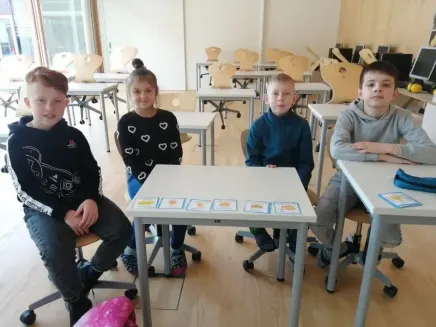 Vier Kinder sitzen an einem Schreibtisch in einem Klassenzimmer und lächeln für ein Foto. Dahinter sind mehrere Tische und Stühle in Reihen angeordnet. Das Klassenzimmer hat eine weiße Wand und einen Holzboden.