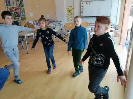 Vier Kinder gehen in einem Klassenzimmer mit Holzböden. Drei von ihnen sind Jungen und eines ist ein Mädchen. Sie tragen Freizeitkleidung. Hinter ihnen stehen Tische und Stühle. An der Wand befindet sich eine Tafel.