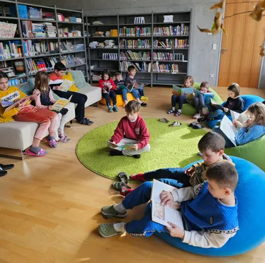 Kinder sitzen auf Sitzsäcken und Sofas in einer Bibliothek. Sie lesen alle Bücher. Die Regale hinter ihnen sind mit Büchern gefüllt.