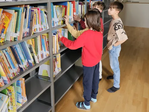 Drei Kinder stehen in einer Bibliothek und schauen sich Bücher auf einem Regal an. Eines trägt Rot, eines Gelb und eines Blau.